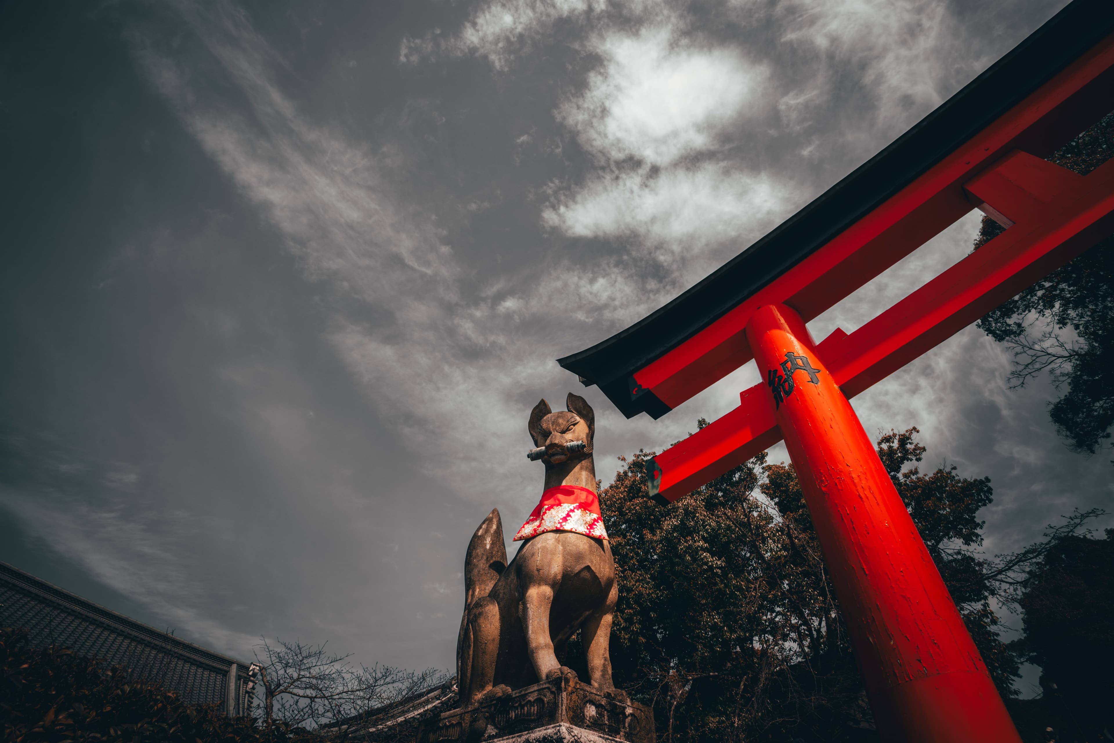 A stone fox guards a red torii gate against a heavy grey sky at a Kyoto shrine.