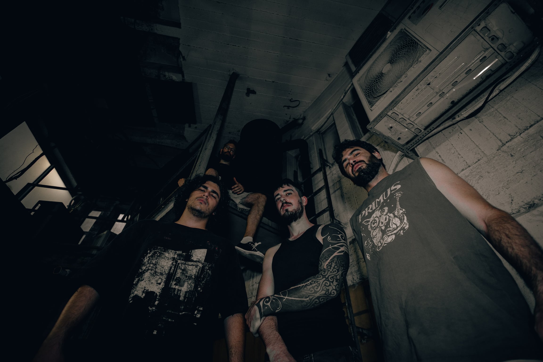 A low-angle shot of the band gathered on a stairwell, one member perched above the others against exposed pipework and an old air conditioning unit.