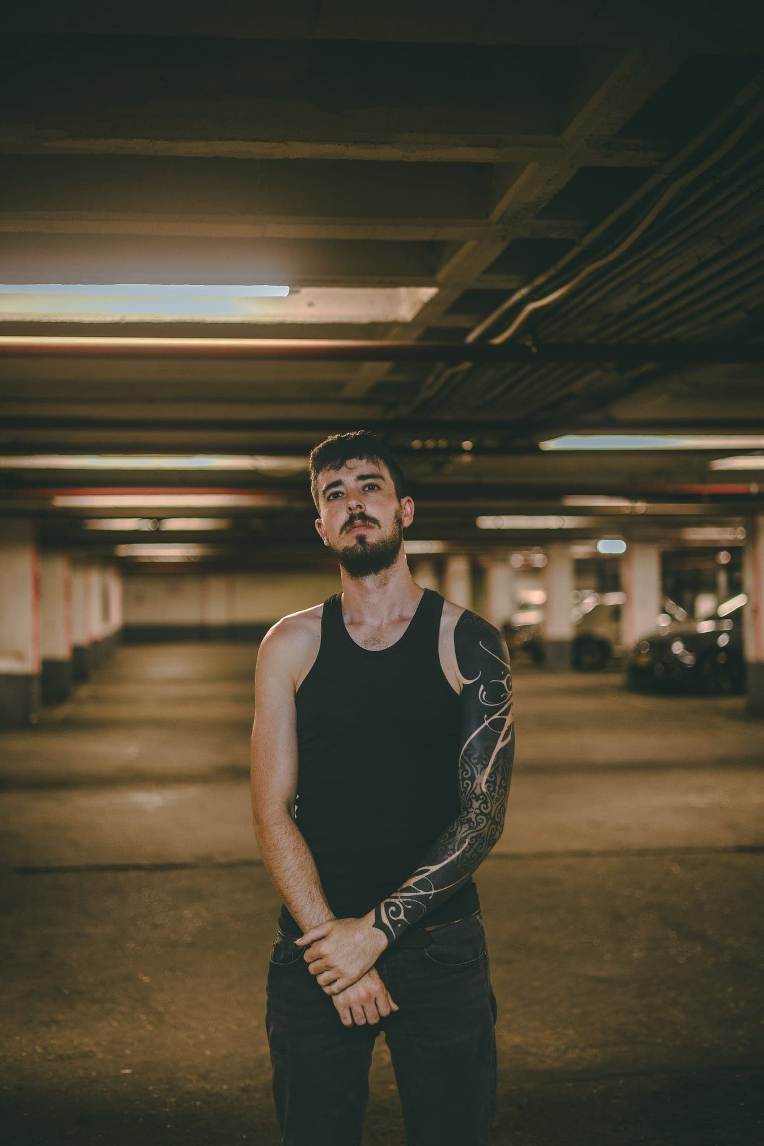 A band member stands alone in an empty parking garage, arms crossed, a full tattoo sleeve visible down one arm.