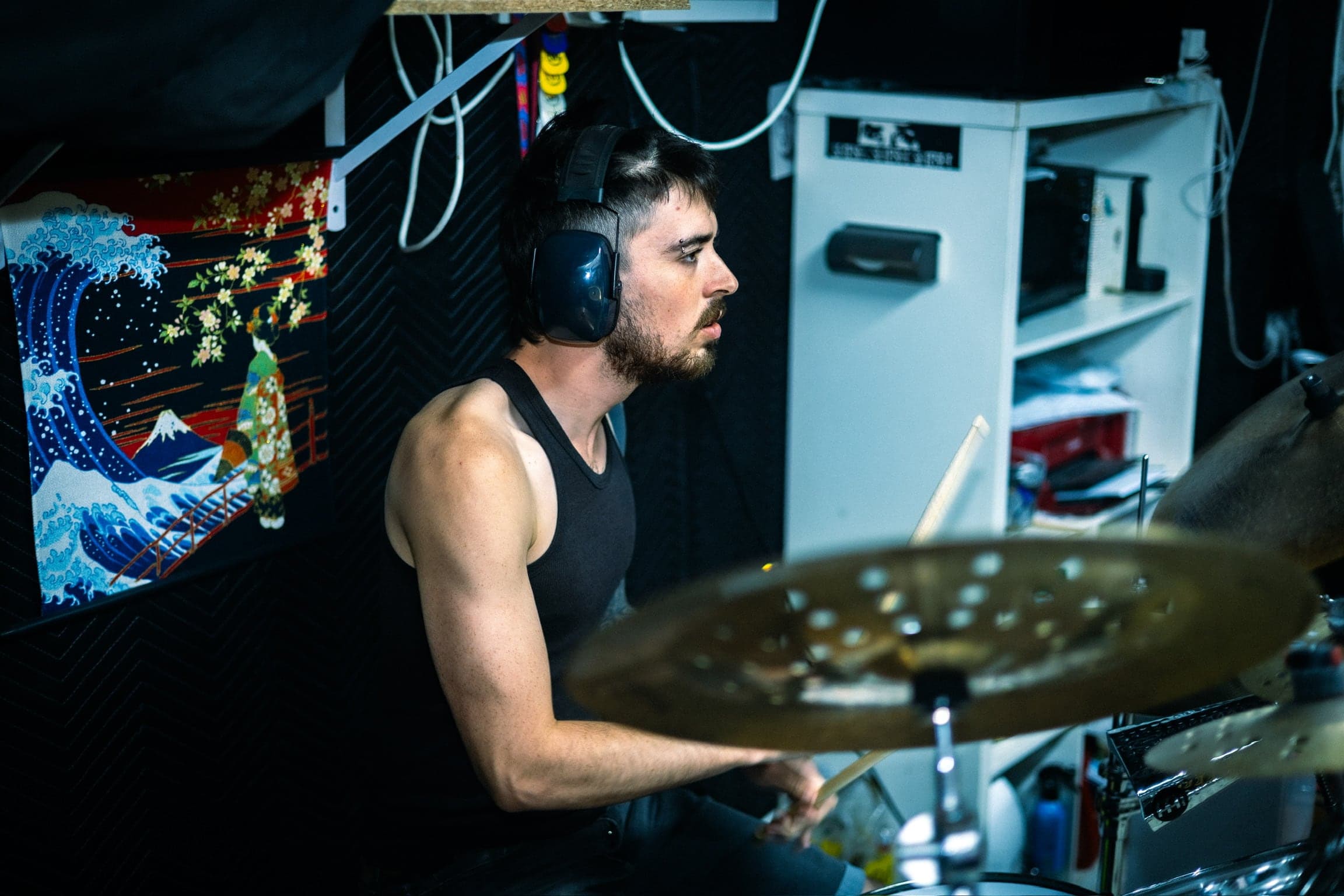 The drummer, headphones on, mid-hit at the kit, a Hokusai wave tapestry pinned to the wall behind him.