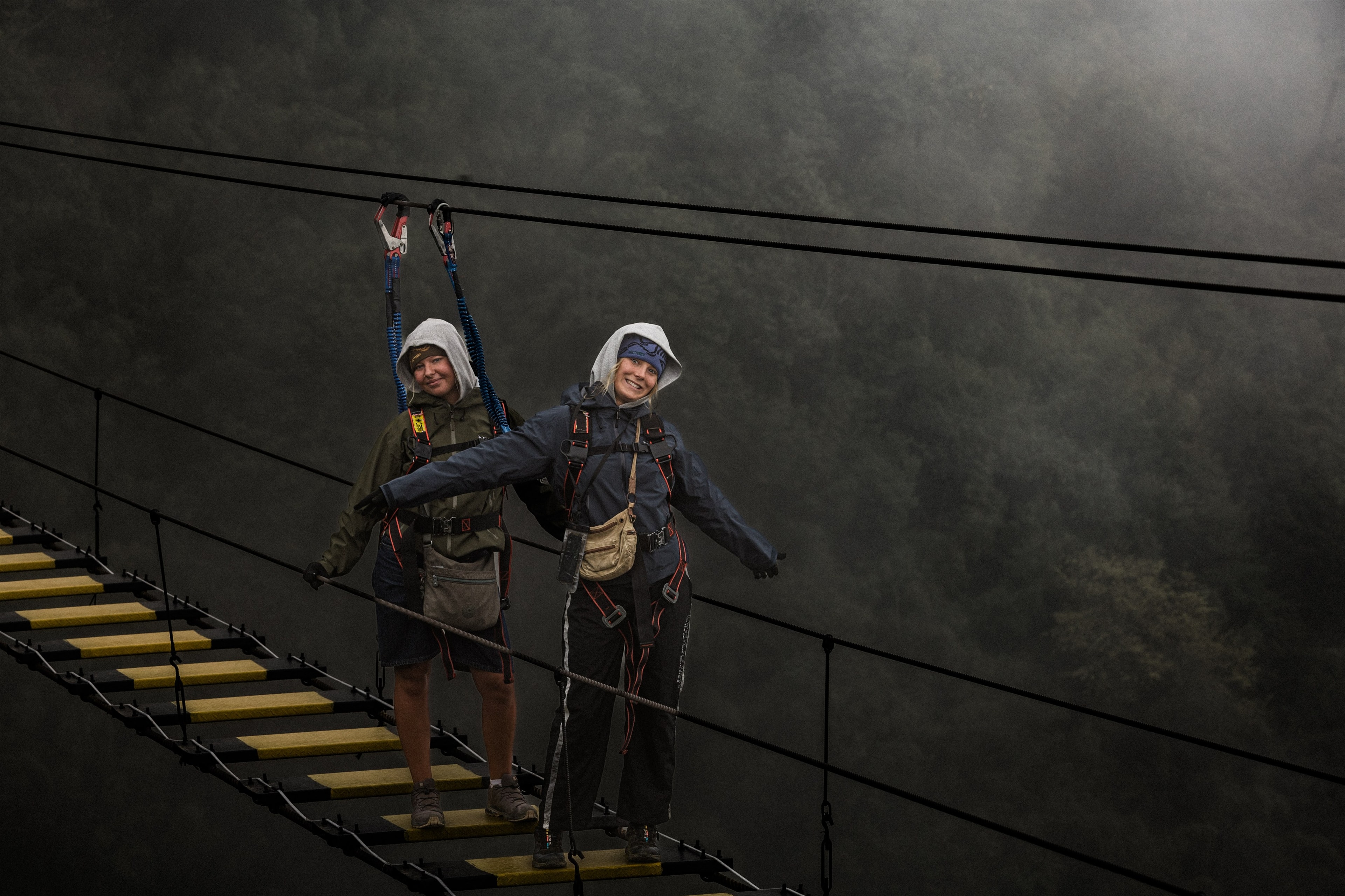 Two climbers clipped into a suspension rope bridge, fog filling the valley behind them.