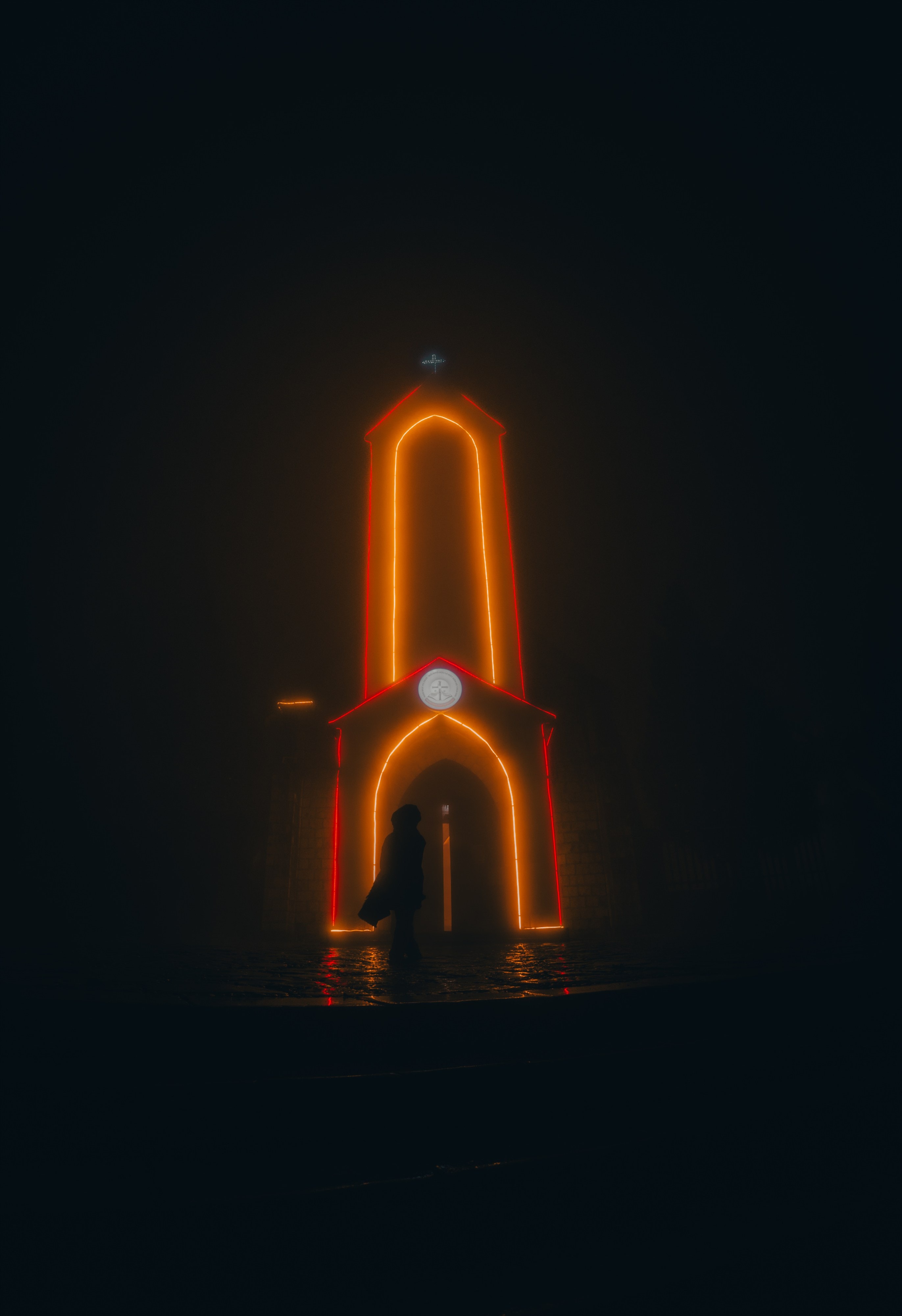 A silhouetted figure standing inside a neon-outlined gate shaped like a church, lit red and gold at night.