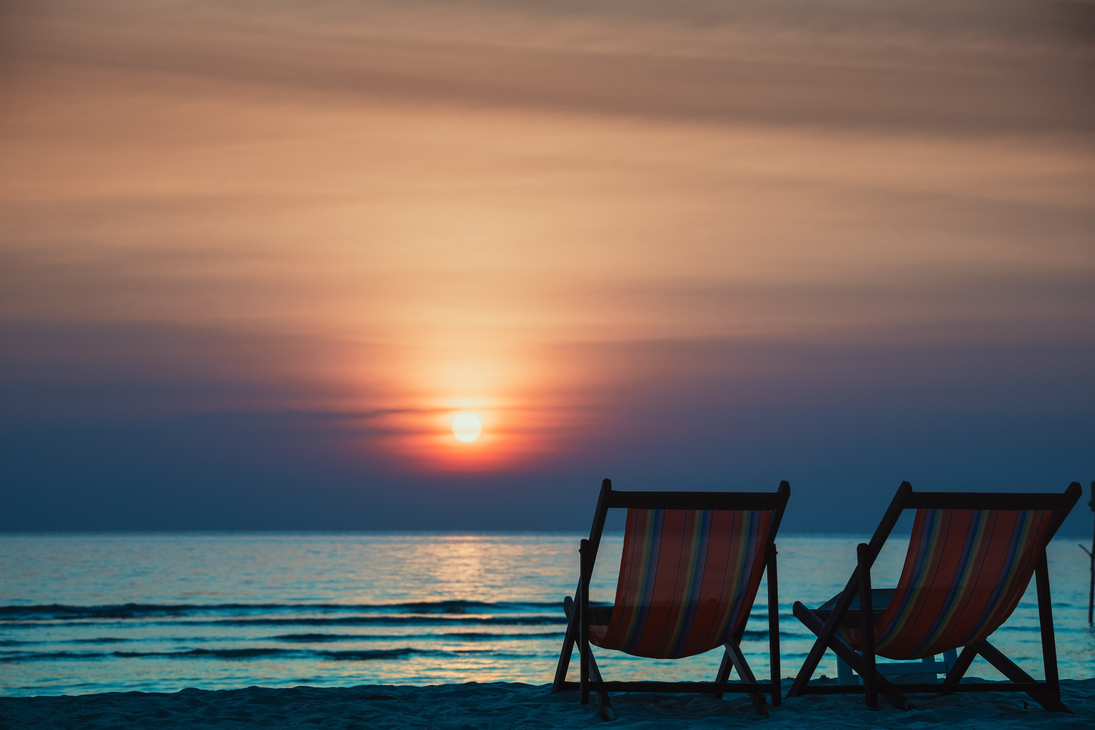 Two empty beach chairs face a burnt-orange sunset over a calm sea.