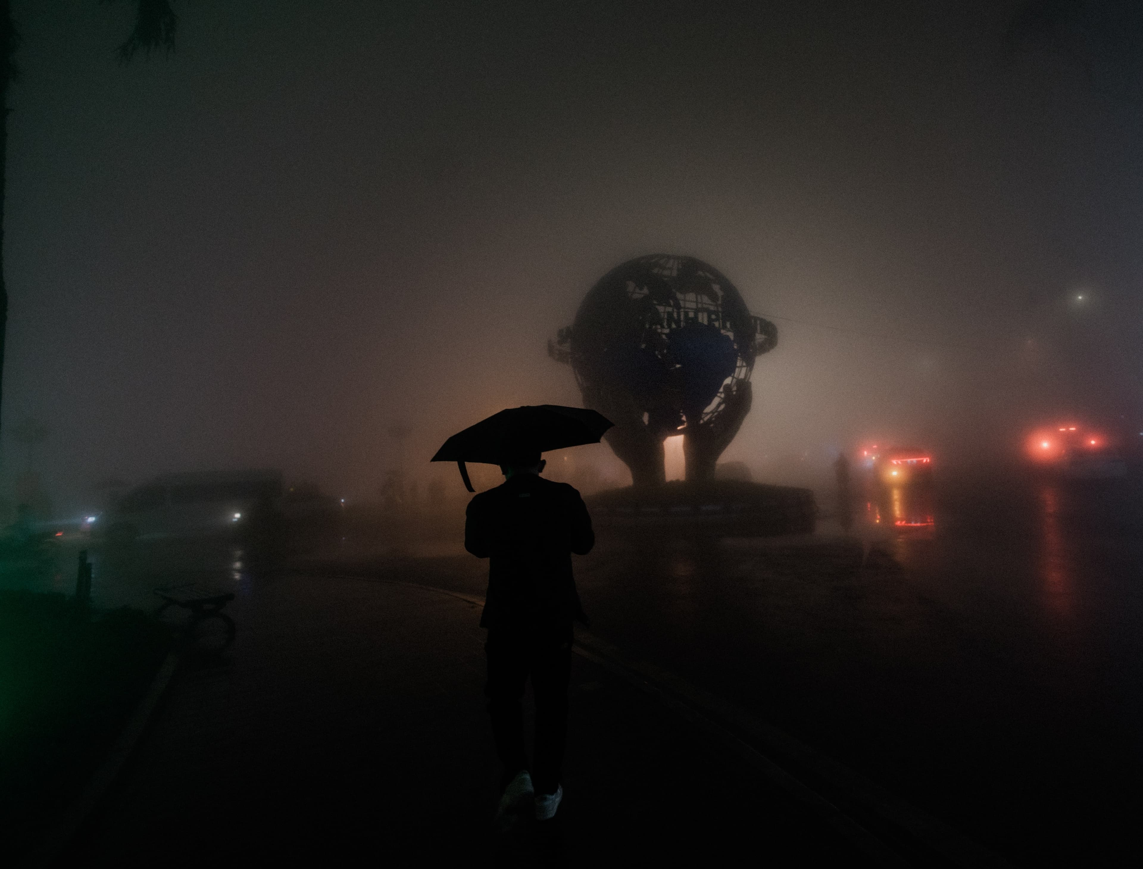 A figure with an umbrella crosses a foggy square at night, a lit globe monument glowing behind them.
